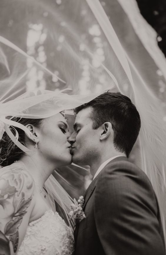 Close-up of a bride and groom sharing a kiss under a flowing veil. The black-and-white tone enhances the intimate and romantic atmosphere. The bride has a visible floral tattoo on her shoulder, and both wear formal wedding attire.