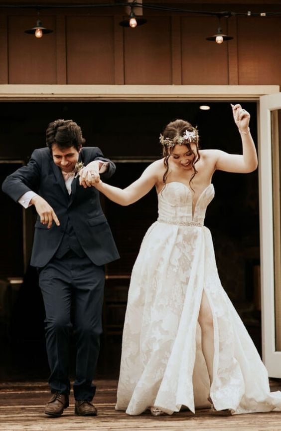 Bride and groom stepping joyfully out of a doorway, holding hands and dancing. The bride wears a strapless, slit wedding gown and a floral crown, while the groom is in a navy suit. The moment is playful and celebratory.