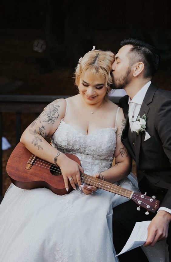 Bride smiling while holding a ukulele as the groom kisses her on the head. They sit close together, radiating affection. Both have visible tattoos and wear classic wedding outfits. The bride has bleached blonde hair styled with clips.
