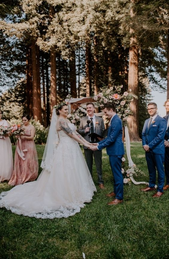 Outdoor wedding ceremony beneath tall redwoods. Bride and groom hold hands in front of a floral arch, with the officiant and wedding party nearby. The setting is natural and elegant, with warm afternoon light filtering through the trees.