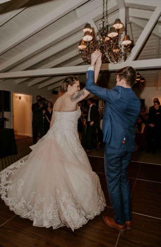 Bride and groom dancing indoors under a chandelier. The bride wears a voluminous lace gown, and the groom is in a blue suit. The bride twirls gracefully while guests watch in the background. The mood is joyful and romantic.