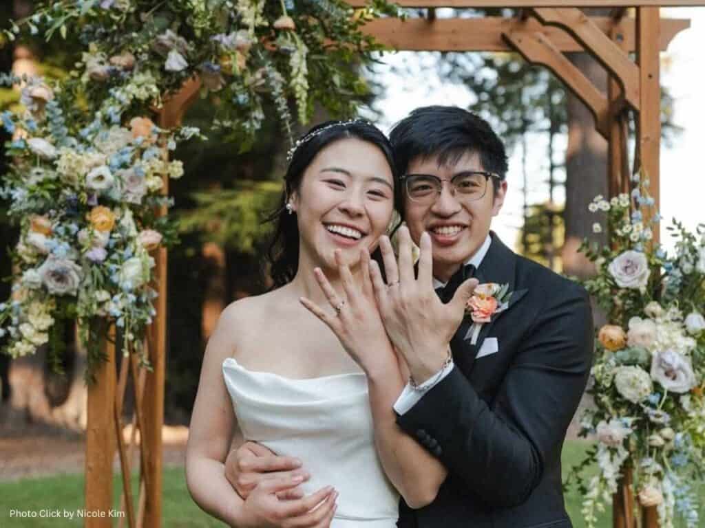 Happy couple showing their wedding rings under a floral arch during an outdoor ceremony