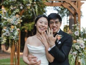 Happy couple showing their wedding rings under a floral arch during an outdoor ceremony