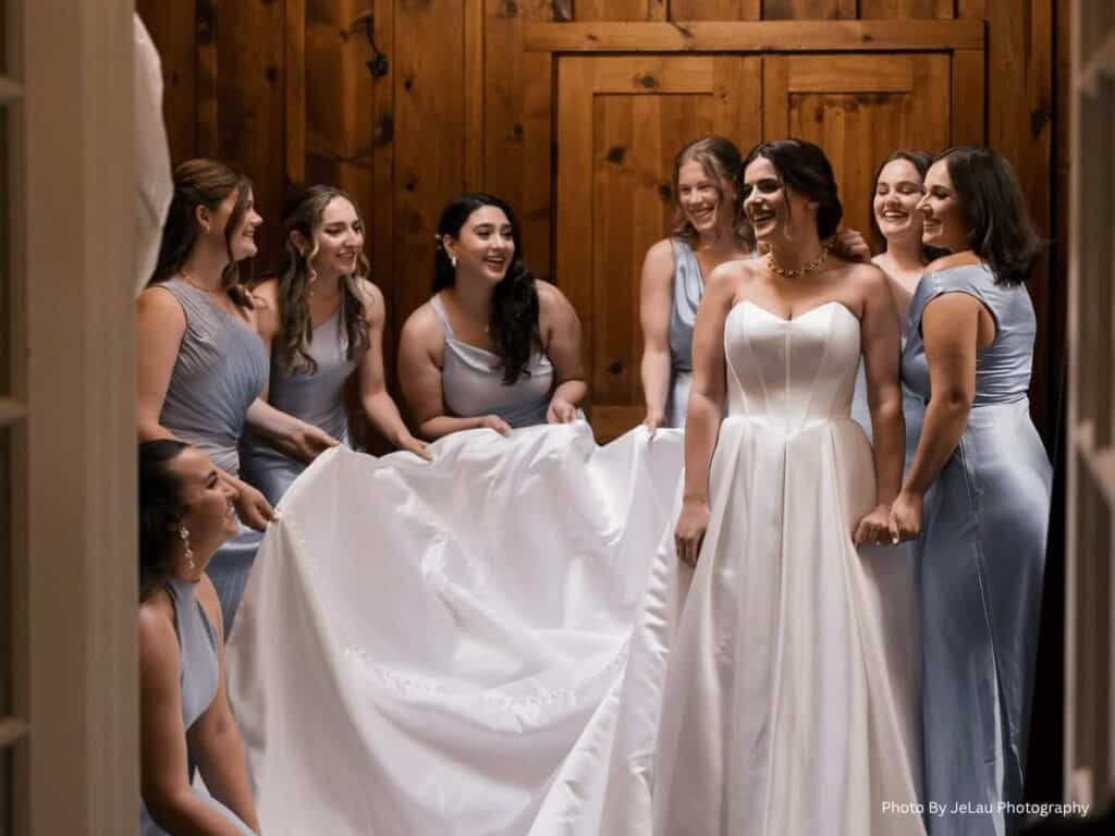 Bride smiles with bridesmaids holding her gown in a wooden room