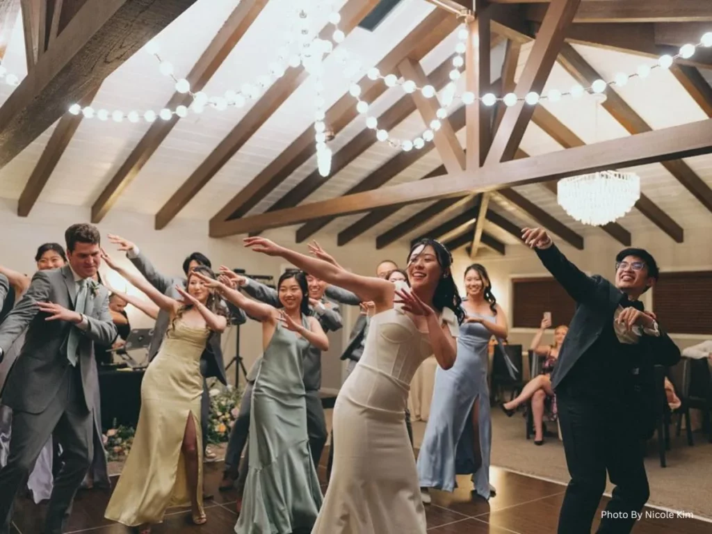 Wedding party dancing together at reception with joyful group celebration and string lights overhead