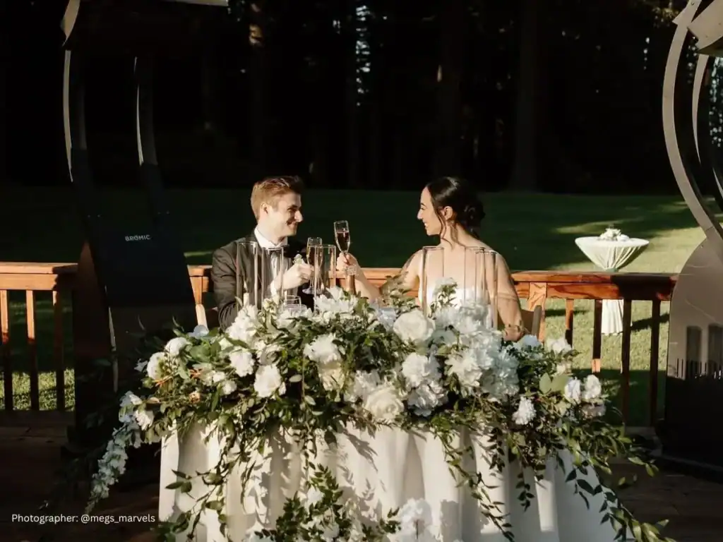 Bride and groom toast at their flower-filled sweetheart table during the wedding reception 